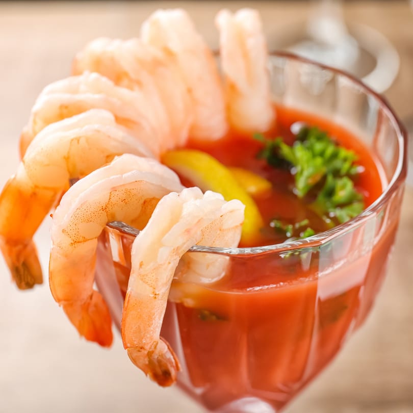 Shrimp cocktail with large shrimp hanging over a glass of red sauce, garnished with lemon and parsley.