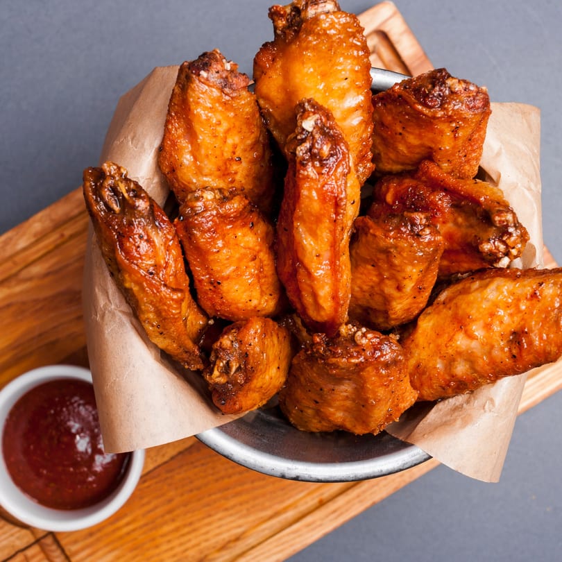 Crispy, golden-brown chicken wings in a metal bowl with parchment paper, served with a side of sauce.